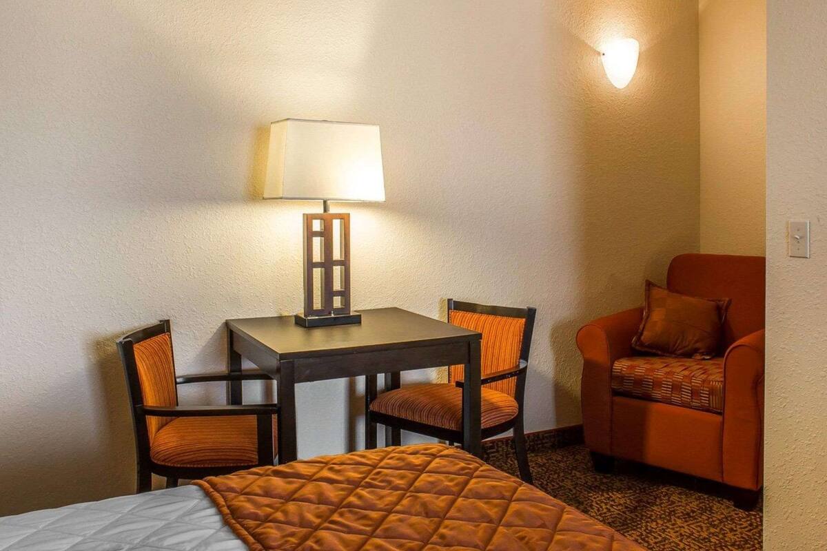 A hotel room with a bed, a table with a lamp, two chairs, and an orange armchair.