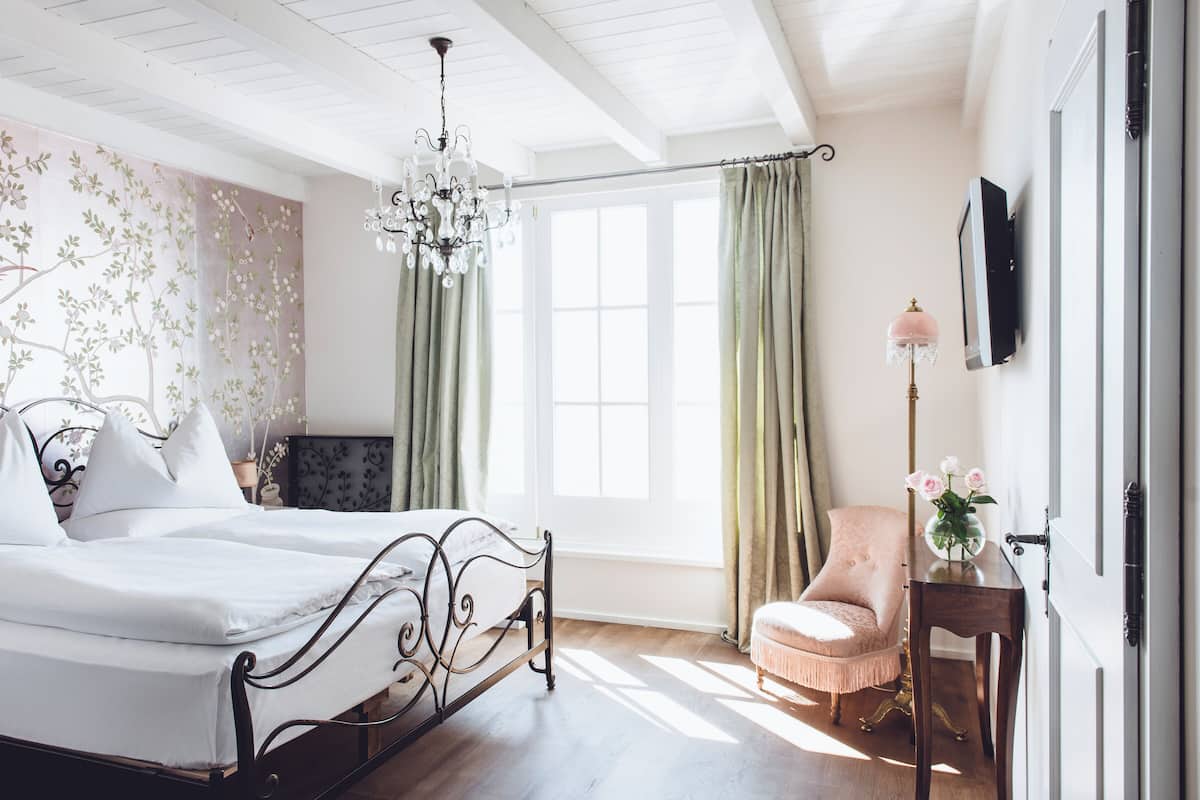 A bedroom with a bed, a chair, a desk with a lamp, a TV, and a window with curtains.