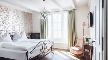 Een slaapkamer met een bed, een stoel, een bureau met een lamp, een televisie en een raam met gordijnen.