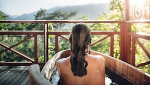 Une personne se détend dans un bain à remous en bois, avec une vue panoramique sur les montagnes et la verdure.