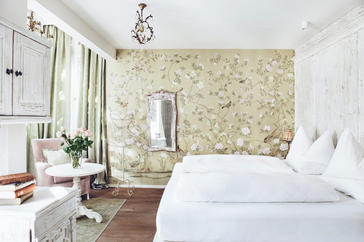 A bedroom with a floral wallpaper, a bed, a chair, a table, and a mirror.