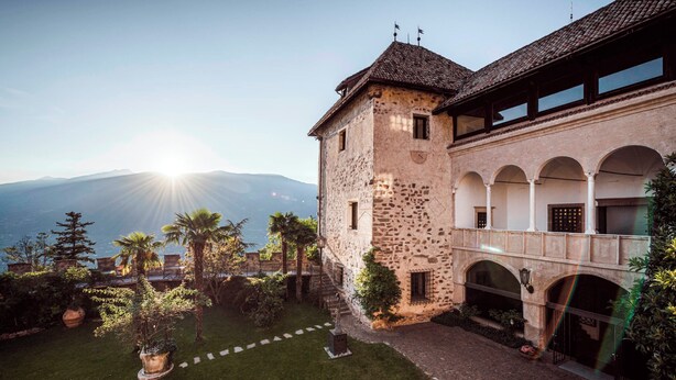 Un bâtiment historique en pierre, doté d’une cour, d’arcades et offrant une vue sur les montagnes en arrière-plan.
