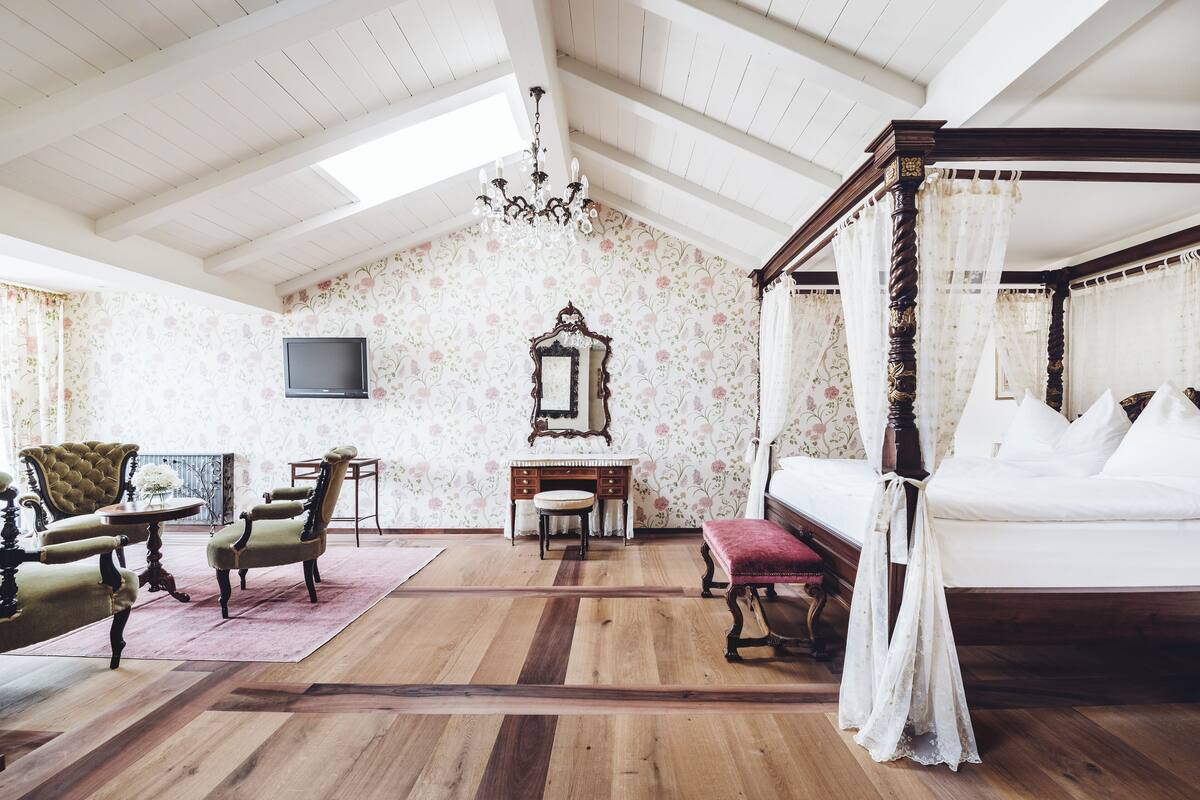 A room with a wooden floor, a four-poster bed, a vanity with a mirror, a television, and floral wallpaper.