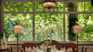 Un coin repas avec une table dressée pour deux personnes, entouré de chaises en bois, d’un lustre et de grandes fenêtres donnant sur un paysage verdoyant.