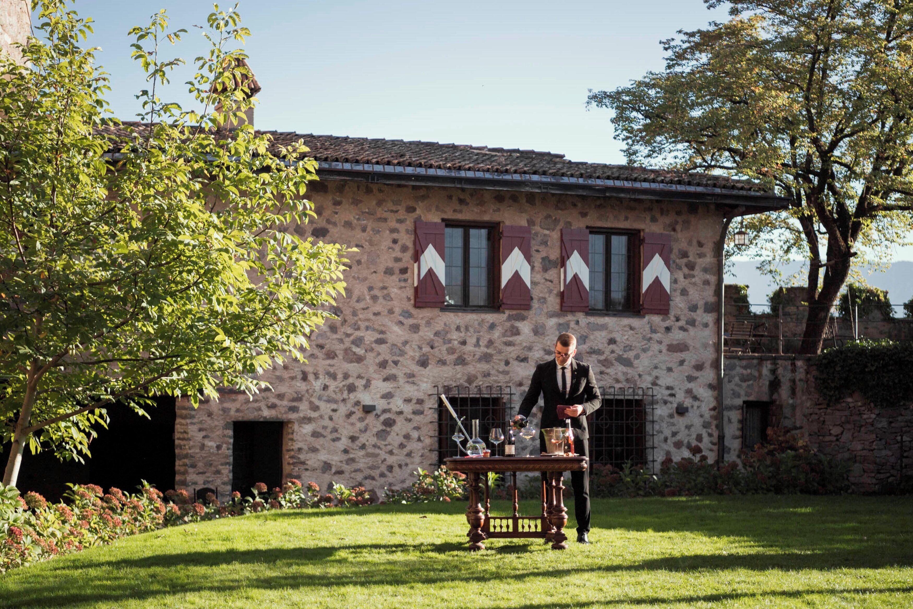 A stone building with a rustic facade, a table with wine glasses and bottles, and a person in a suit standing beside it.