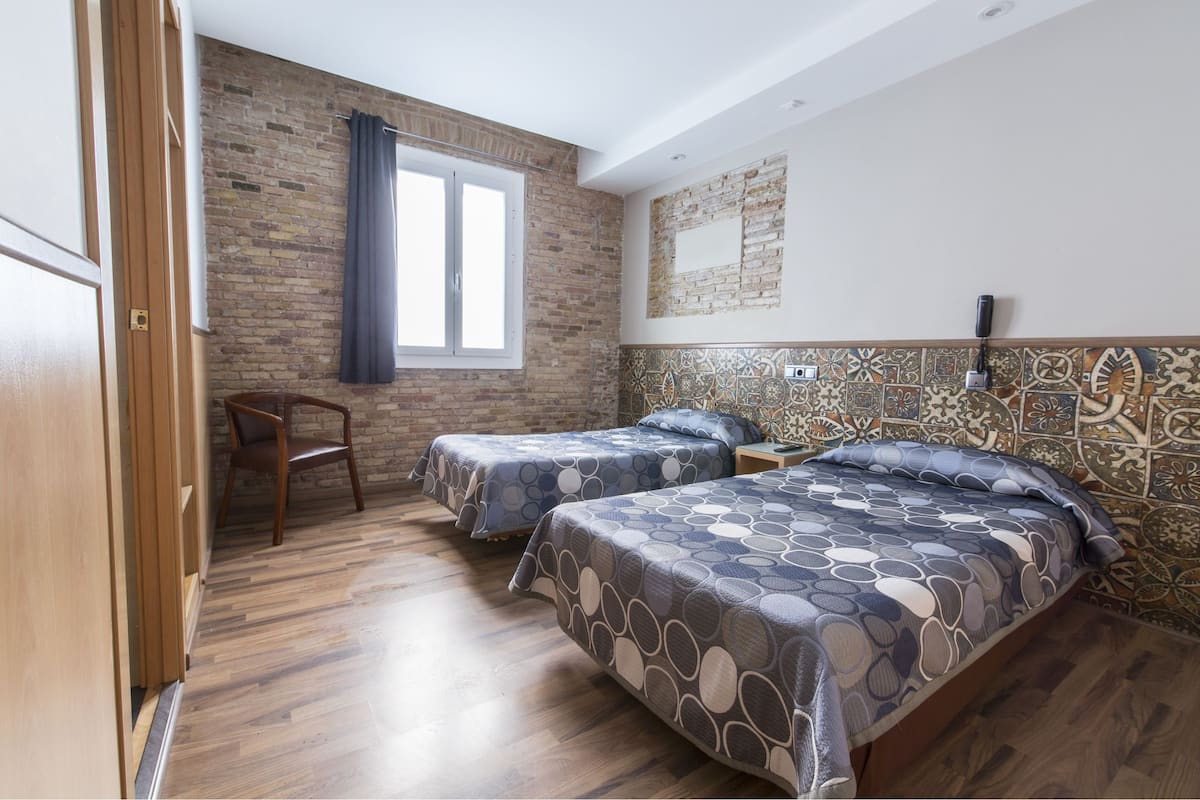 Quarto com duas camas, uma cadeira de madeira, uma janela e uma parede de tijolos.