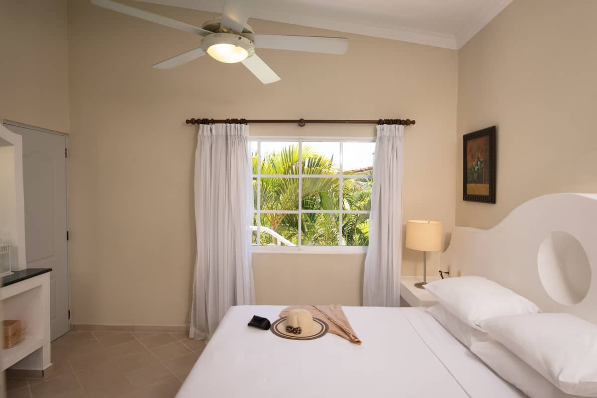 Ein Hotelzimmer mit Bett, Nachttisch, Lampe, Deckenventilator und einem Fenster mit Vorhängen.