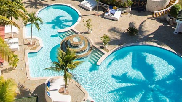 A large outdoor swimming pool with a circular water feature, surrounded by lounge chairs and palm trees. - Lifestyle Crown Residence Suites - All Inclusive (Puerto Plata)
