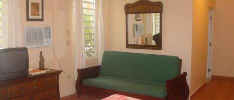 A room with a bed, a green sofa, a wooden dresser, a television, a mirror, and a window with curtains.
