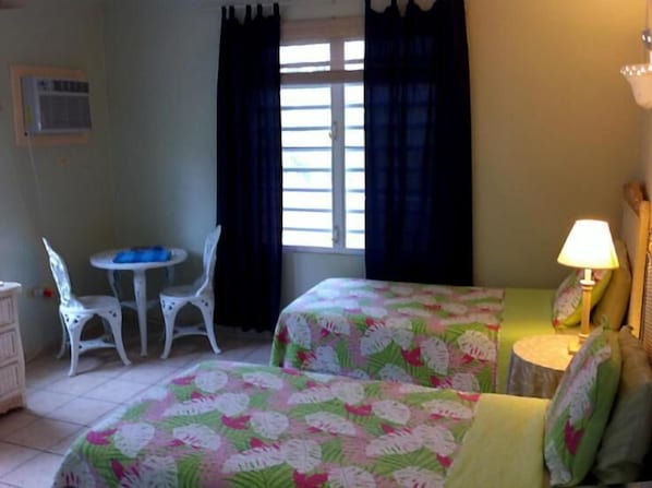 A room with two beds, a small table, two chairs, a lamp, and a window with curtains. - Dos Angeles del Mar B&B (Rincon)