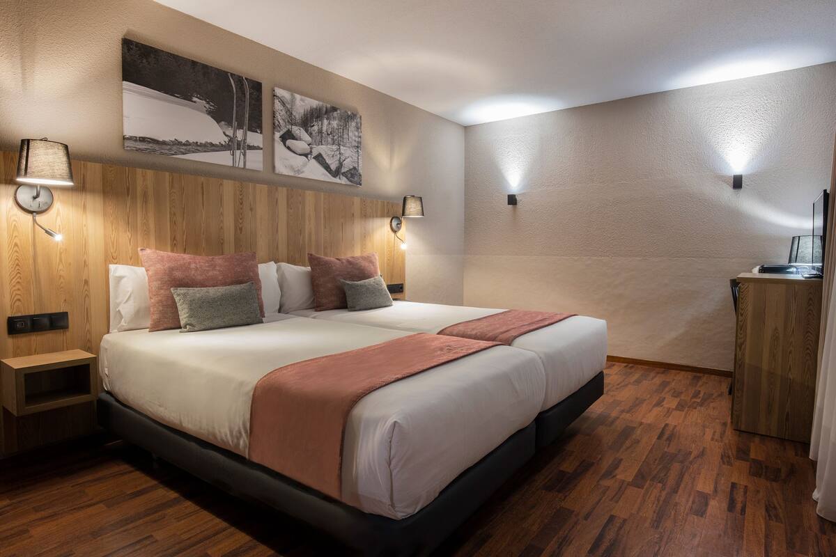 A modern hotel room with a large bed, wooden headboard, two bedside tables, wall-mounted lamps, and a painting above the bed.