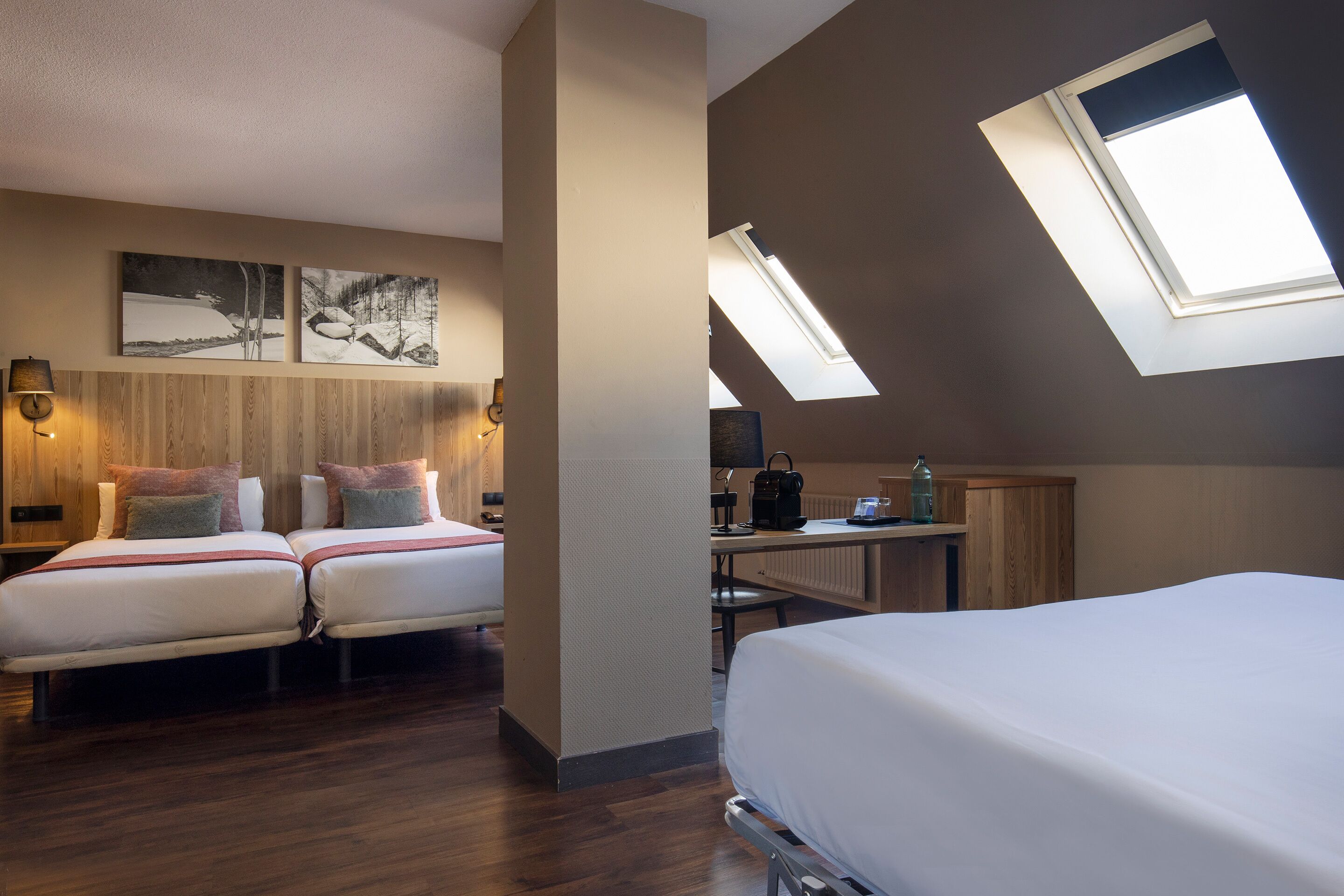 A hotel room with two beds, a desk, a radiator, and a skylight.