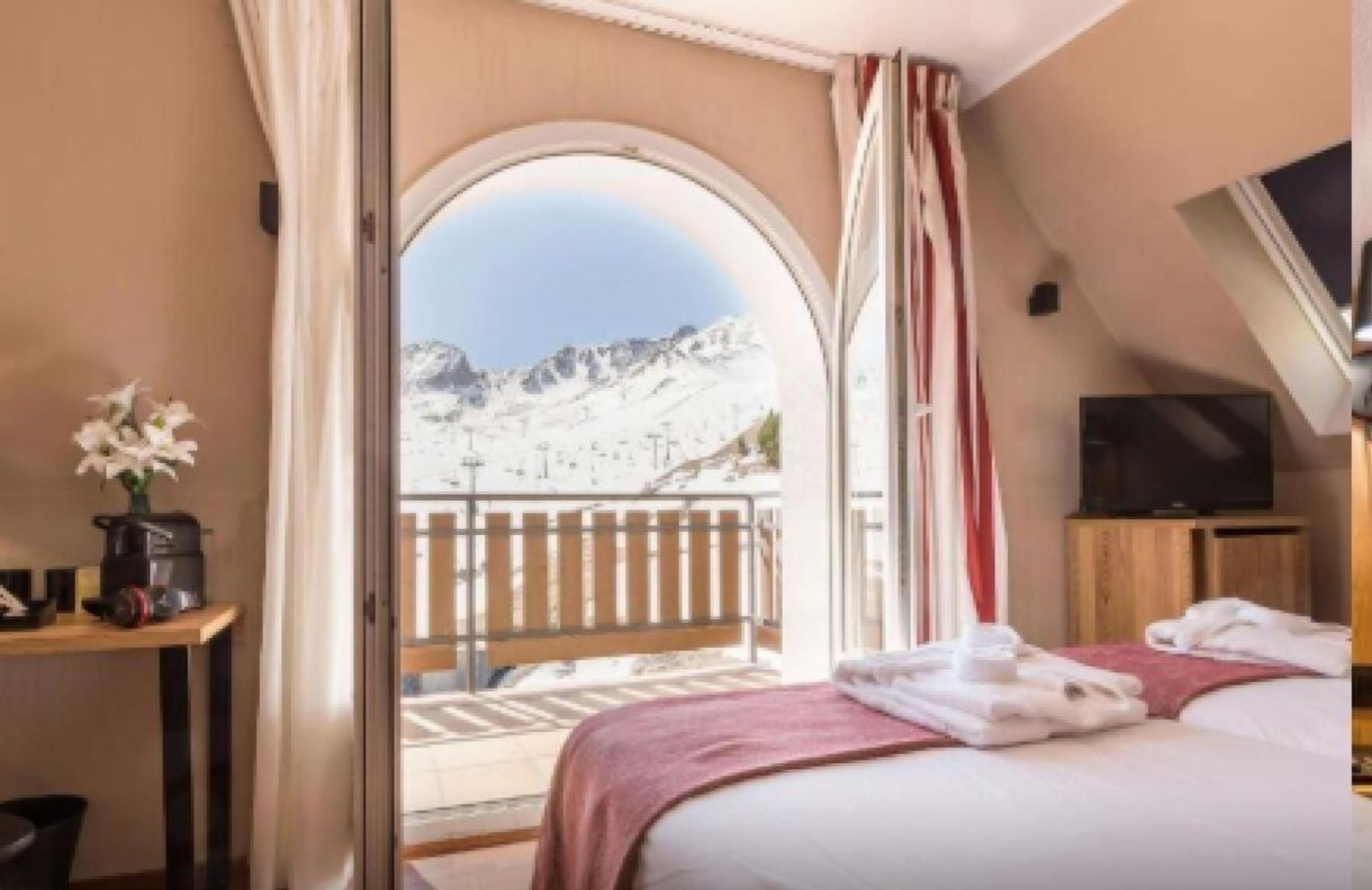 Une chambre d’hôtel avec un lit, une télévision, une table agrémentée d’un vase de fleurs et un balcon offrant une vue sur la montagne.