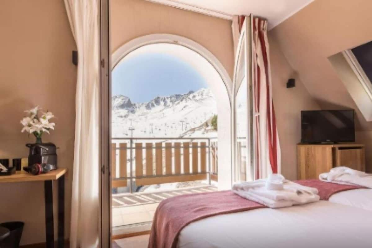 Une chambre d’hôtel avec un lit, une télévision, une table agrémentée d’un vase de fleurs et un balcon offrant une vue sur la montagne.