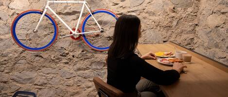 A person sitting at a wooden table with a meal, a bicycle hanging against a stone wall, and a chair in the foreground.