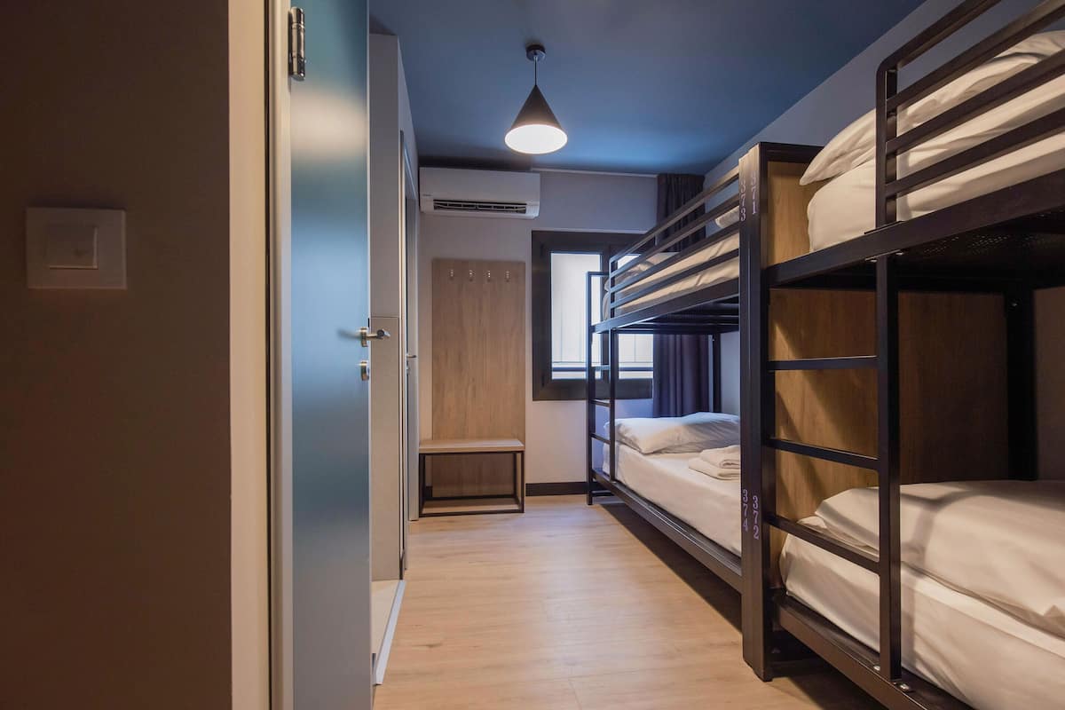 Habitación de hotel compacta con literas, un banco pequeño y una ventana que deja entrar luz natural.