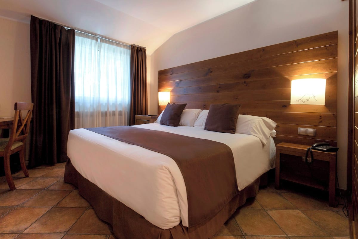 Une chambre d’hôtel avec un grand lit, une tête de lit en bois, deux lampes de chevet, une table de chevet en bois, une chaise et une fenêtre avec des rideaux.