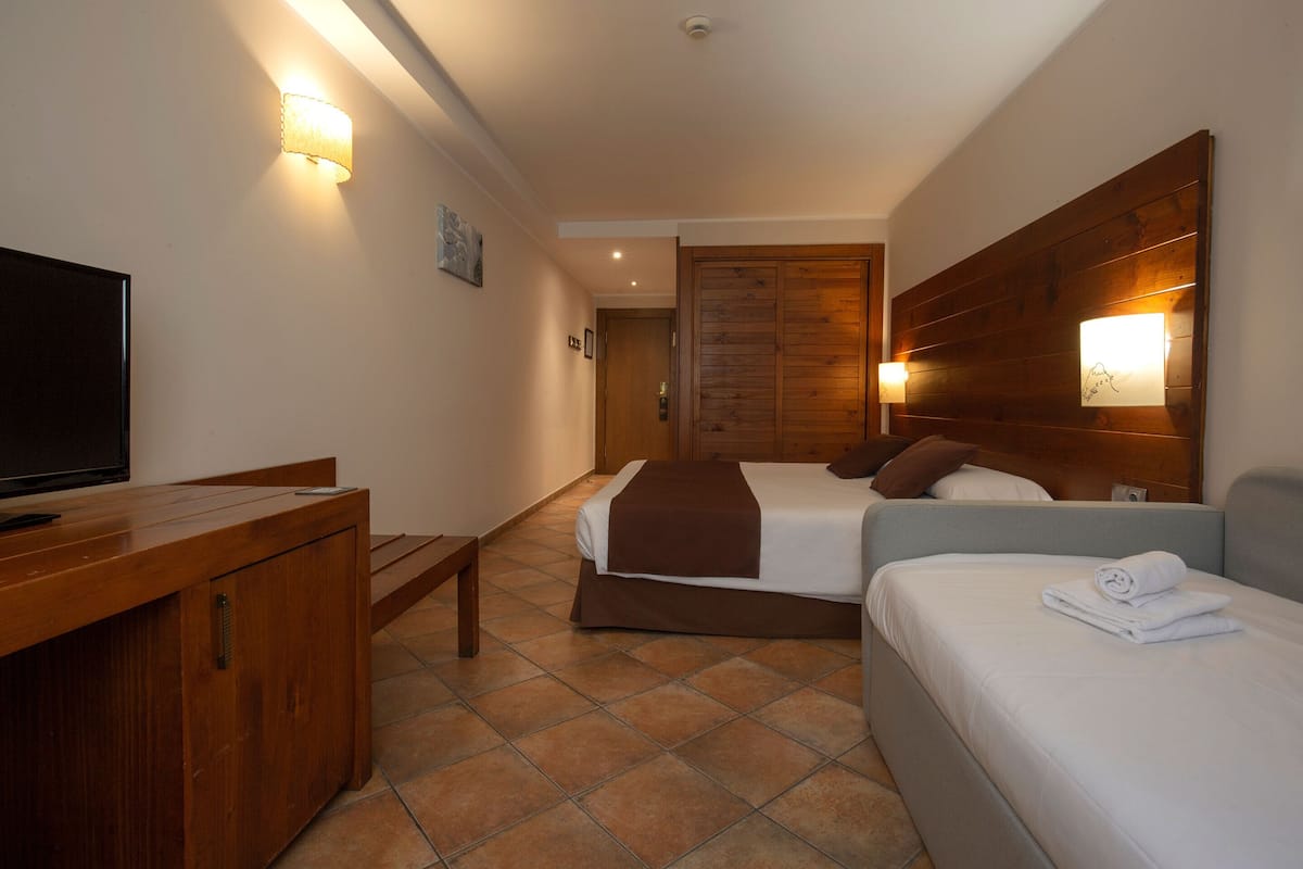 Une chambre d’hôtel avec un bureau en bois, une télévision, un lit recouvert d’une couverture marron et une table de chevet avec une lampe.
