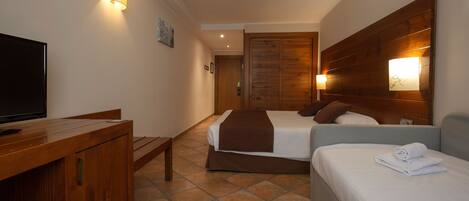 A hotel room with a wooden desk, a television, a bed with a brown blanket, and a bedside table with a lamp.