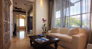 A spacious living room with a beige sofa, a dark wooden coffee table, and a potted orchid. A bookshelf and a door leading to another room are visible.