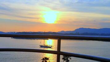 Un coucher de soleil sur une étendue d'eau calme, avec un voilier, un pont et des montagnes au loin.