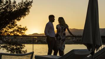Un couple se tient près d’une piscine, avec une vue sur le coucher de soleil, des chaises longues et un parasol.
