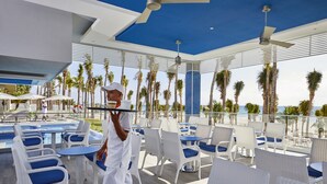 A poolside area with white wicker chairs and blue cushions, a pool, and palm trees in the background. - Hotel Riu Palace Riviera Maya - All Inclusive (Playa del Carmen)