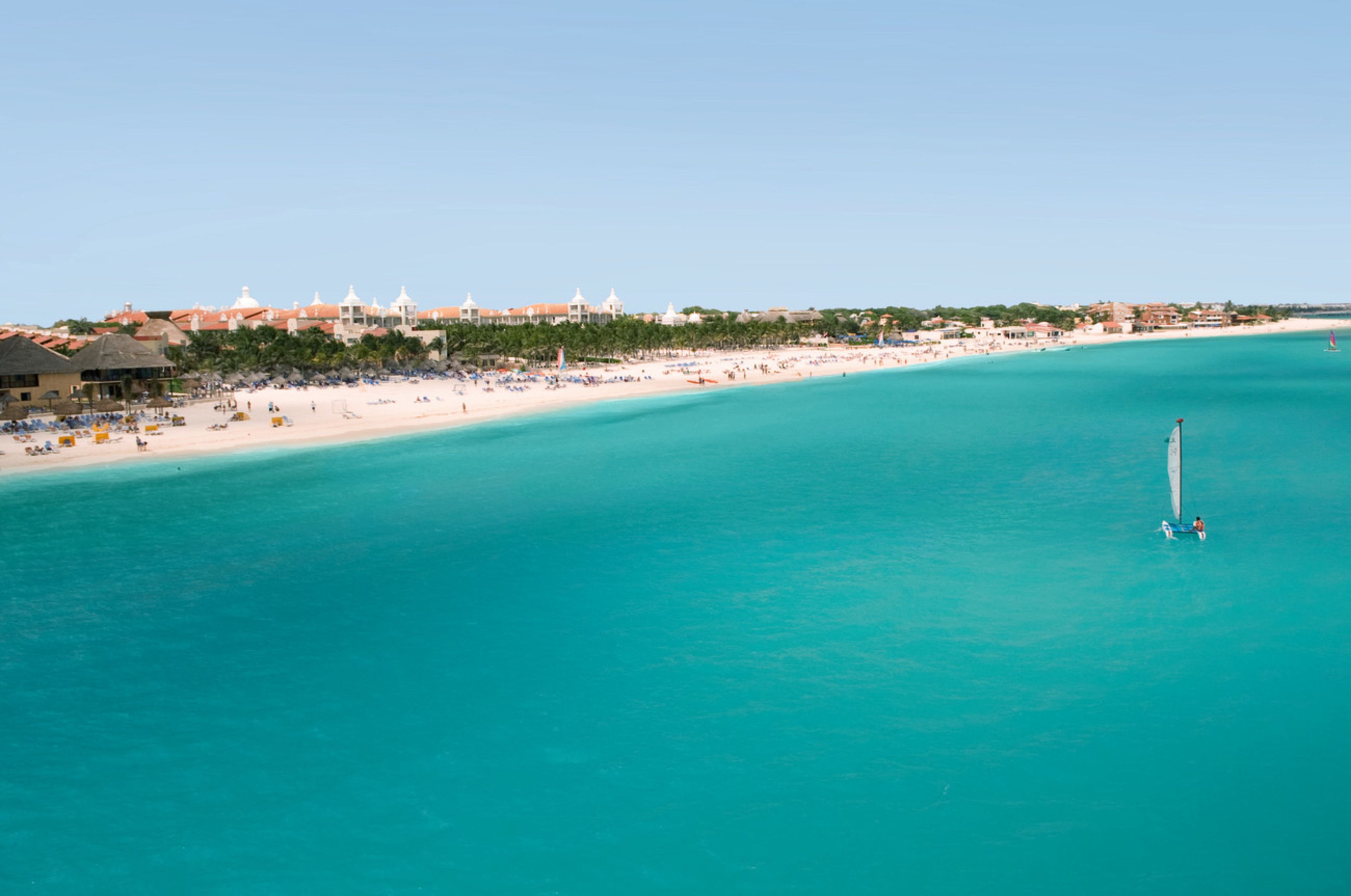 Een kustresort met een zandstrand, helder turquoise water en een zeilboot.