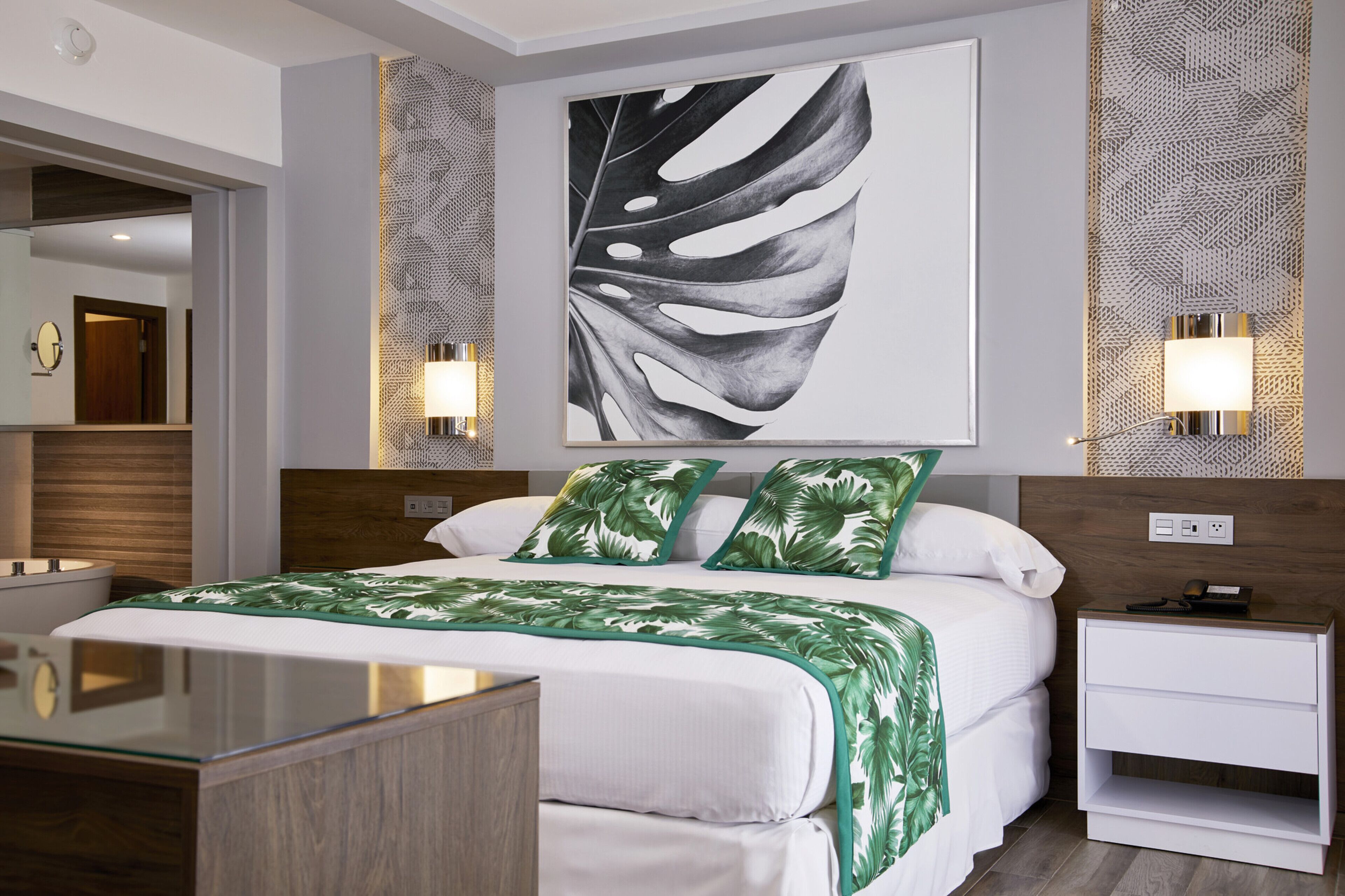 A modern hotel room with a large bed, a bedside table, a nightstand, a mirror, and a wall-mounted artwork.