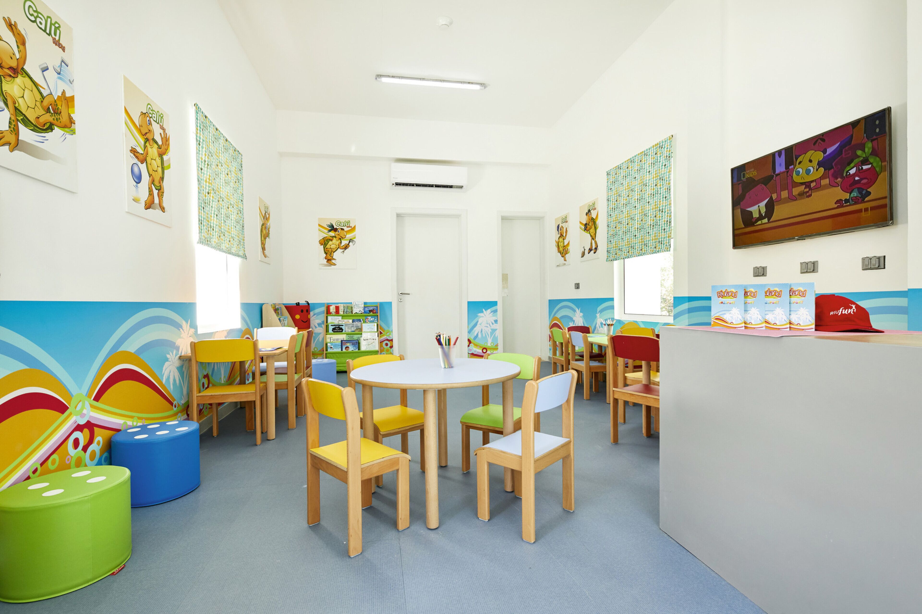 Una sala de juegos infantil muy colorida con una mesa redonda, sillas de colores y carteles educativos en las paredes.