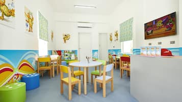 Una sala de juegos infantil muy colorida con una mesa redonda, sillas de colores y carteles educativos en las paredes.
