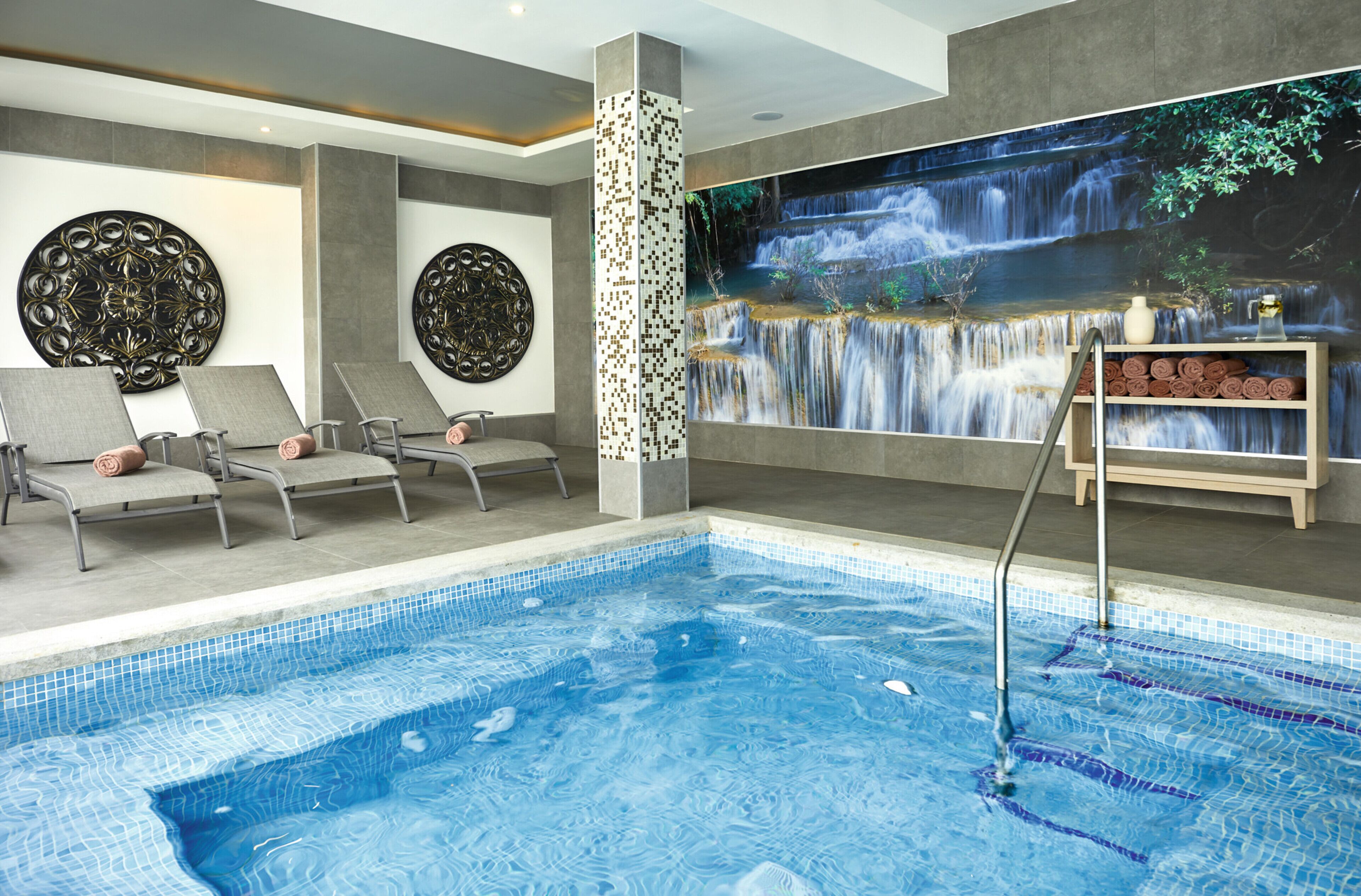 Una piscina interior moderna con un mural de cascada, sillas de descanso y un estante con toallas.