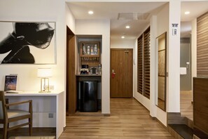 A hotel corridor with a wooden floor, a desk with a chair, a refrigerator, a cabinet with bottles, and a wooden door.