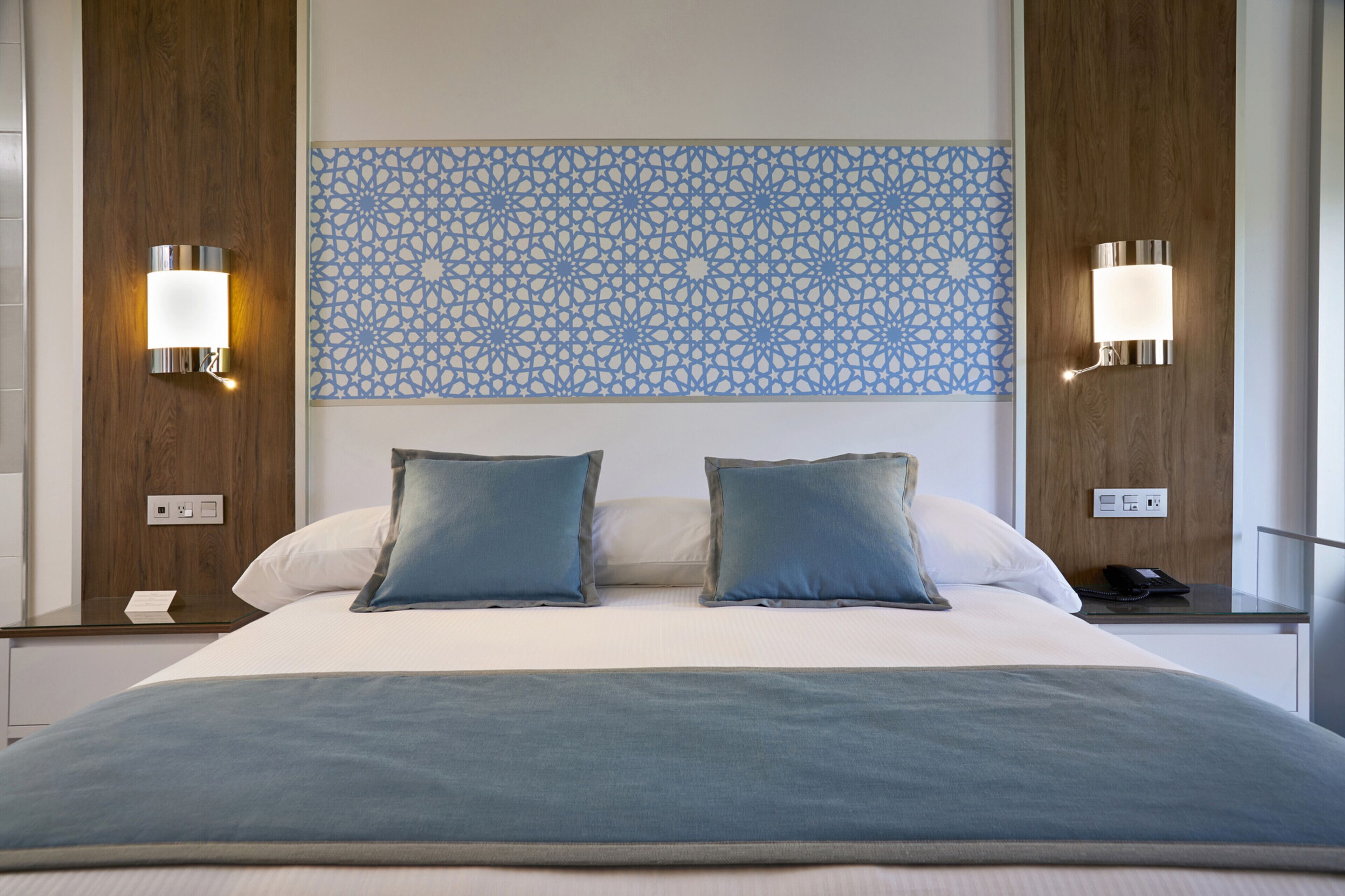 A hotel room with a large bed, two bedside lamps, a decorative wall panel, and a nightstand with a phone and a book.