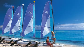 Una playa con varios veleros alineados en la arena, una persona sentada en uno de los barcos y otra de pie en un barco cercano.