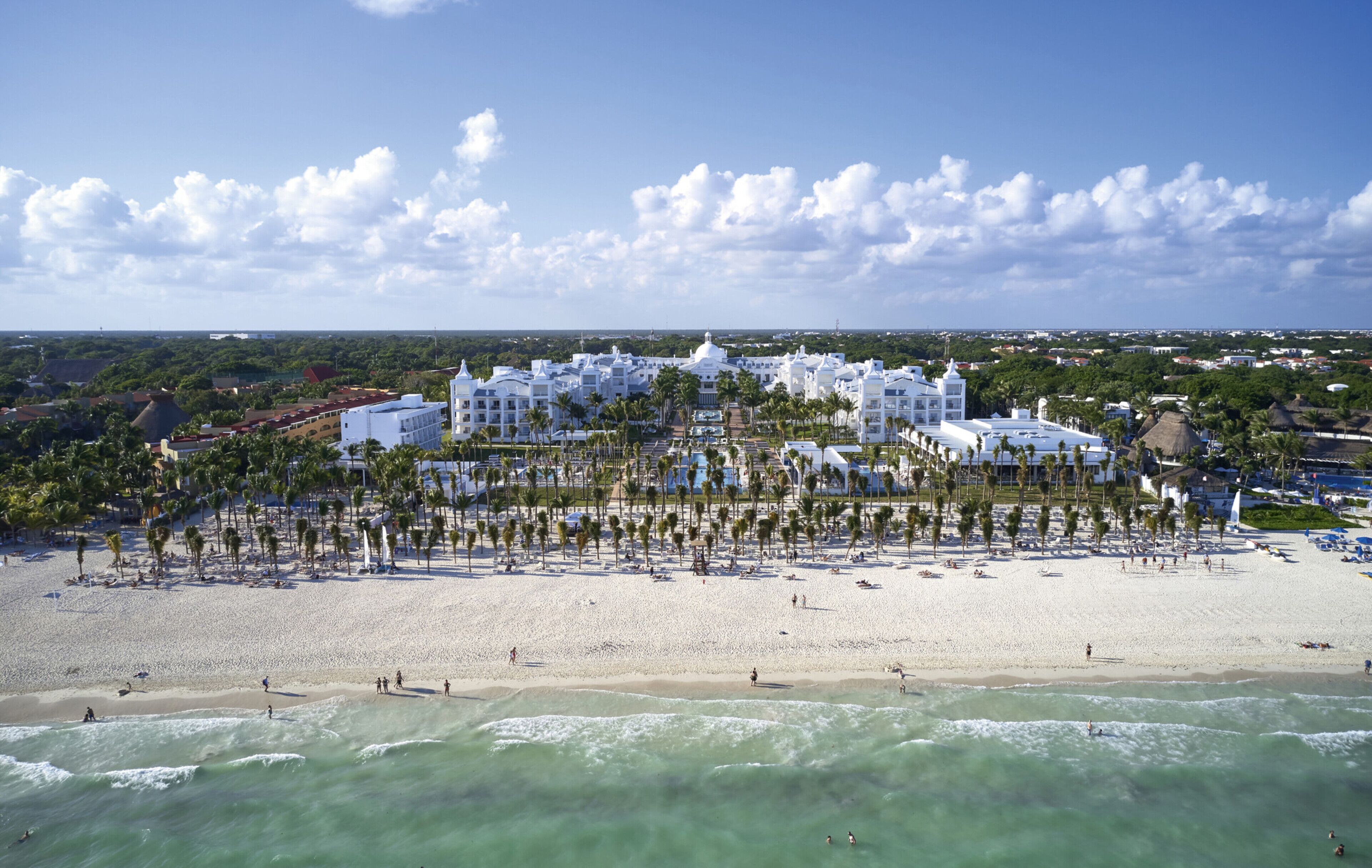 Een hotel aan het strand met meerdere witte gebouwen, een zandstrand en palmbomen.
