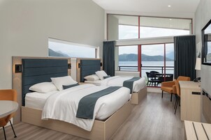 A modern hotel room with two beds, a desk, and a view of the sea.