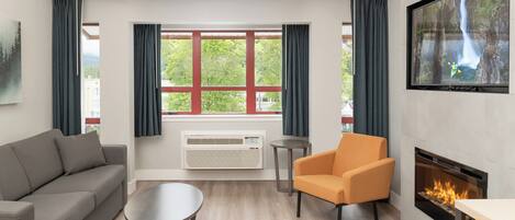 Een moderne woonkamer met een grijze bank, een oranje fauteuil, een klein rond tafeltje, een open haard en een groot raam met gordijnen.