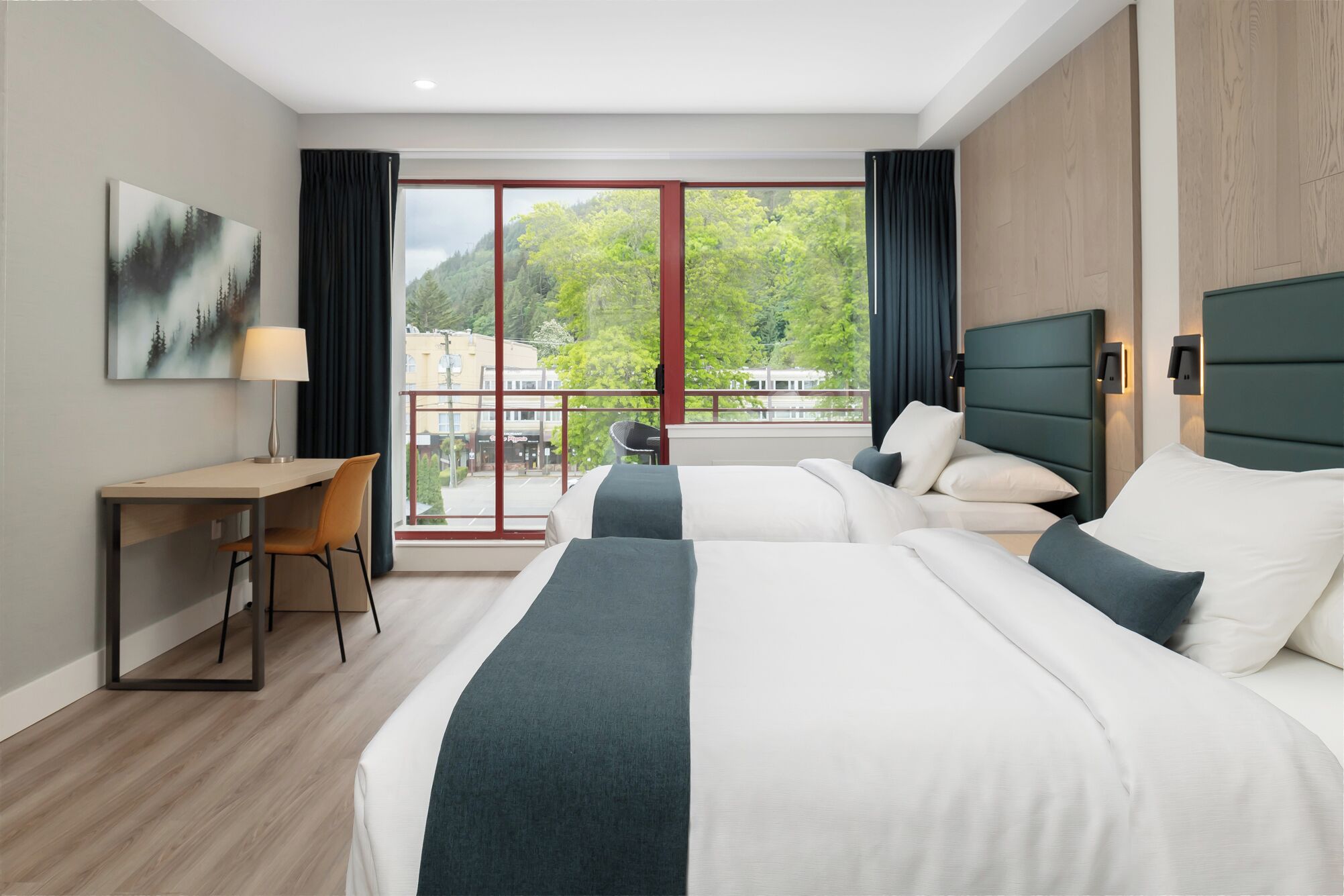 Habitación de hotel con dos camas, un escritorio y vistas a árboles y edificios a través de ventanas amplias.