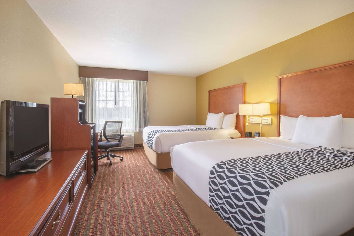 A hotel room with two beds, a desk with a TV, a chair, and a window with curtains.