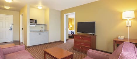 A hotel room with a kitchenette, a flat-screen TV, a sofa, a coffee table, and a lamp.