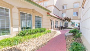A courtyard with a red brick pathway, surrounded by greenery and a building with large windows and a tiled roof. - La Quinta Inn & Suites by Wyndham Bentonville (Bentonville)