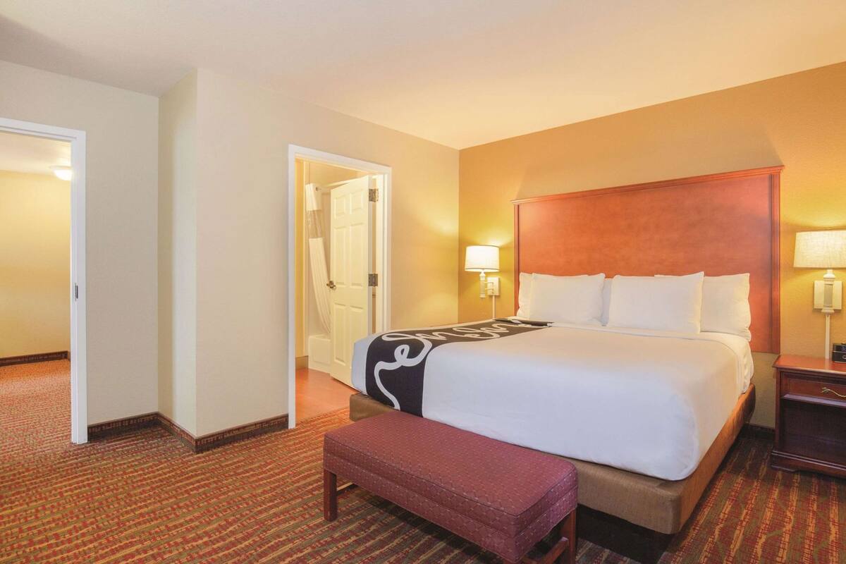 A hotel room with a large bed, a bench, a door, and a nightstand with a lamp.