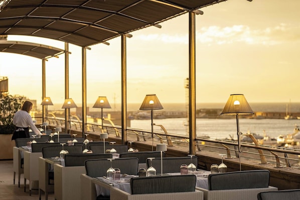 A restaurant with outdoor seating overlooking a waterfront, featuring tables with lamps and a view of the sea.