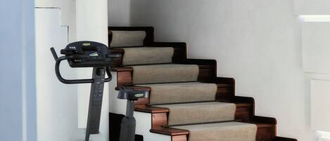 A home gym with a stationary bike and a treadmill, a staircase with beige carpeted steps, and a light fixture on the ceiling.