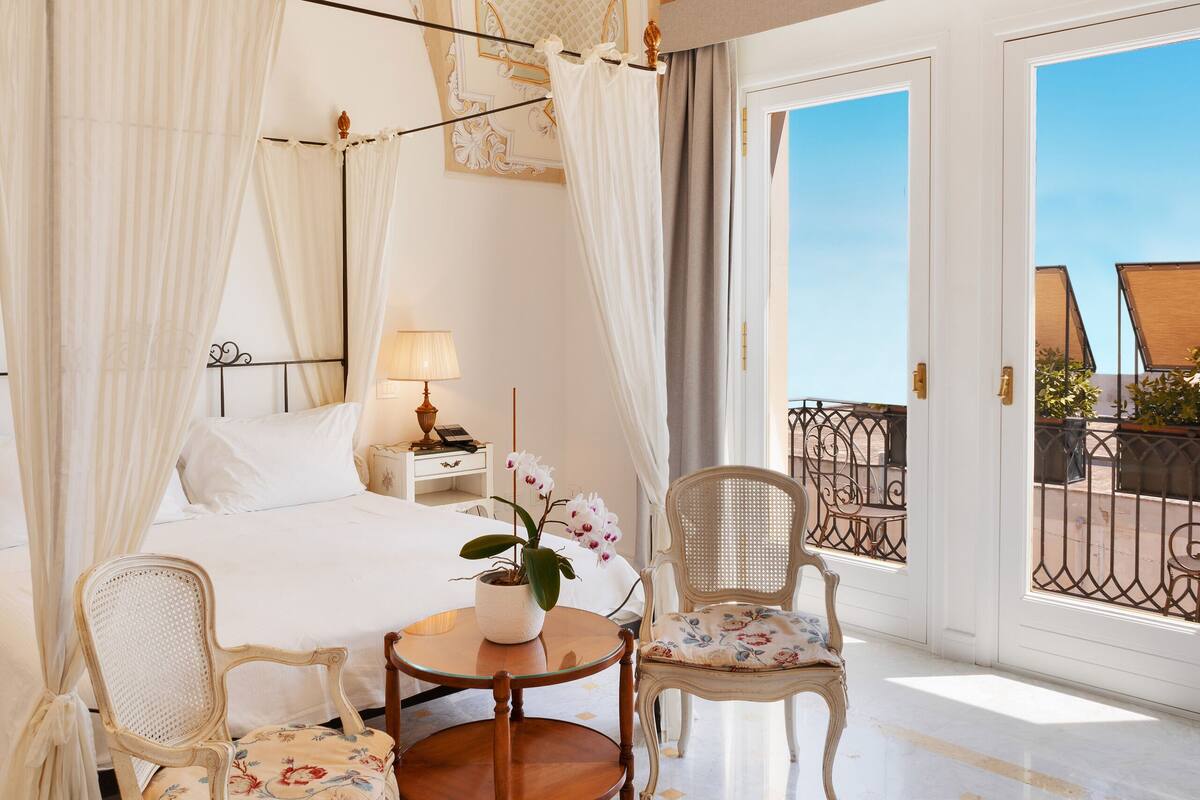 A luxurious bedroom with a four-poster bed, a sitting area with chairs, a small table, and a balcony with outdoor furniture.