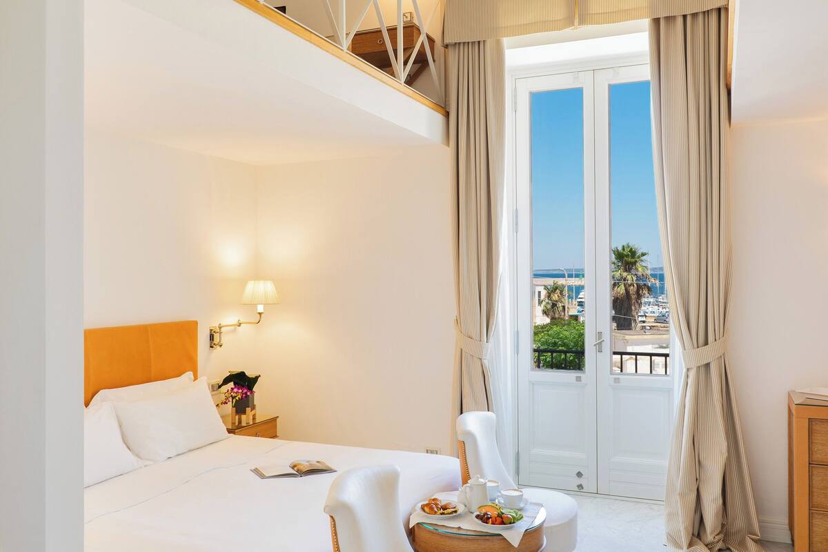 A hotel room with a bed, a desk, a chair, a balcony with curtains, and a view of the sea.