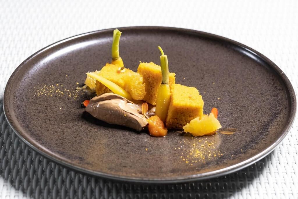 Een donkere keramieken schaal met een dessert bestaande uit gele cake, een stuk chocolade en een klein sinaasappelschijfje.