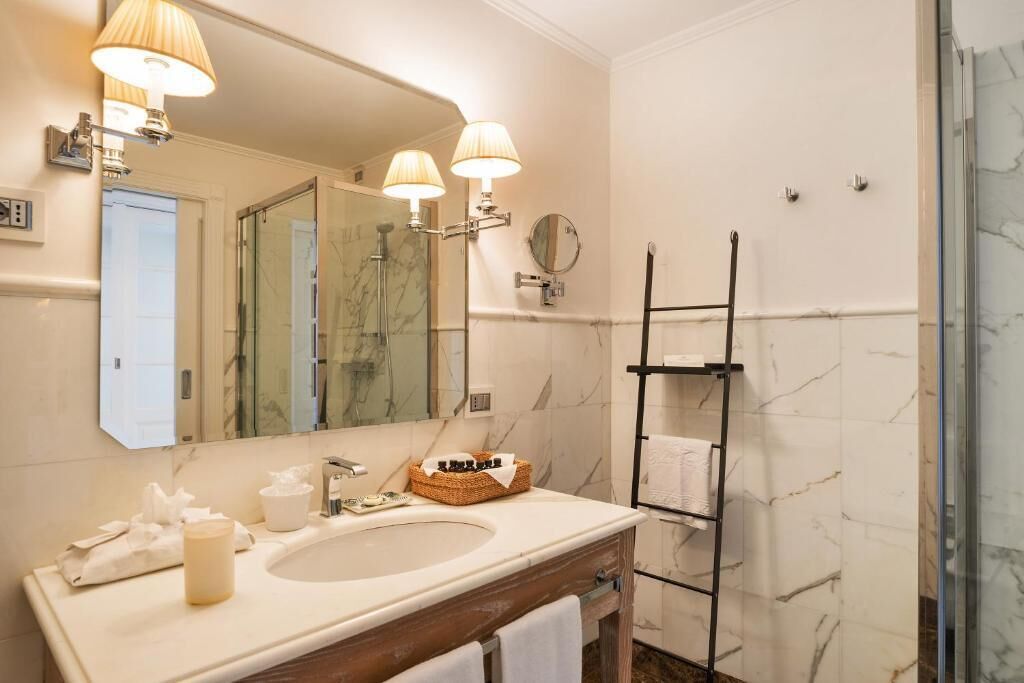 A bathroom with a large mirror, two wall-mounted lamps, a sink, a towel rack, and a shower area.