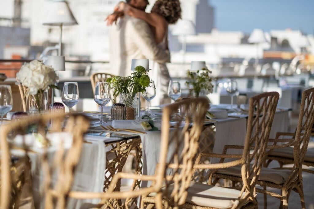 Un área de comedor en la azotea con mesas preparadas para dos, con sillas de mimbre y manteles blancos.